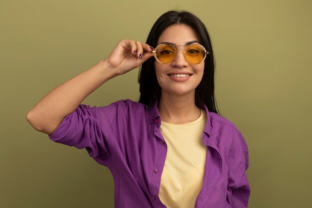 La ragazza caucasica abbastanza mora sorridente in occhiali da sole esamina la macchina fotografica su verde oliva