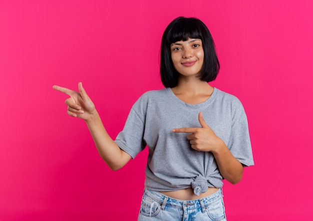 La ragazza caucasica castana giovane lieta indica a lato con due mani che esaminano macchina fotografica isolata su fondo rosa con lo spazio della copia