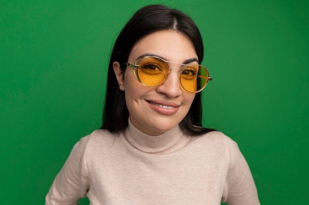 La ragazza caucasica del brunette grazioso sorridente in occhiali da sole esamina la macchina fotografica sul verde