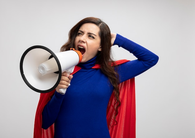 La ragazza caucasica infastidita del supereroe con il mantello rosso mette la mano sulla testa e grida nell'altoparlante guardando il lato isolato sul muro bianco con lo spazio della copia