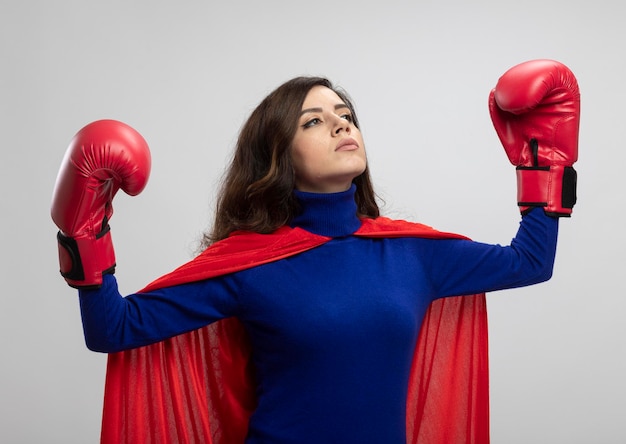 La ragazza caucasica sicura del supereroe con il mantello rosso che indossa i guantoni da pugile si leva in piedi con sollevato