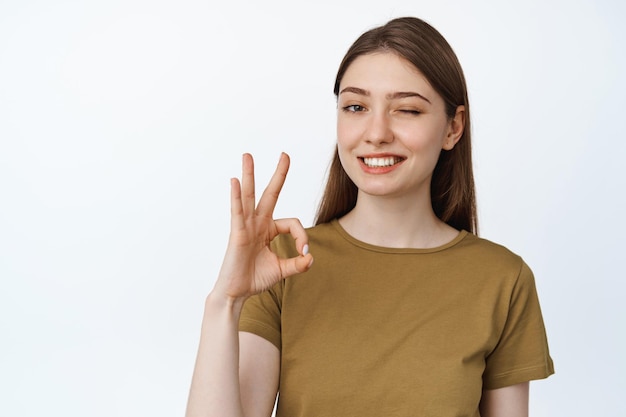 La ragazza che fa l'occhiolino mostra bene, segno OK e sorridente, annuisce in segno di approvazione, consiglia qualcosa di buono, complimenti di qualità, sfondo bianco