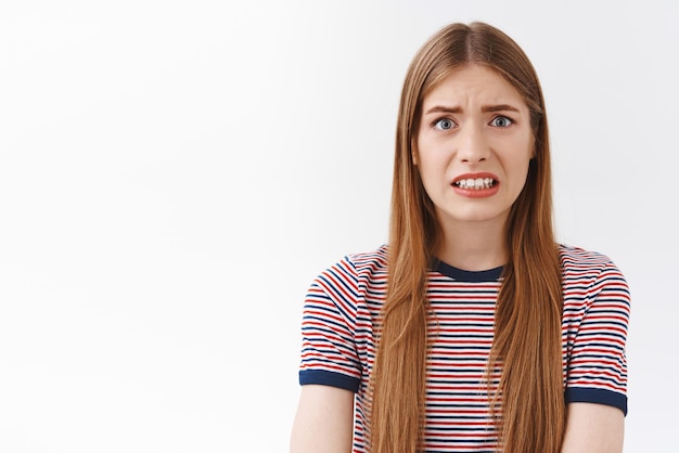 La ragazza che ha paura delle conseguenze sembra imbarazzata e nervosa rabbrividire il viso e stringere i denti accigliata infastidita in piedi preoccupata e ansiosa sfondo bianco ascolta notizie terribili