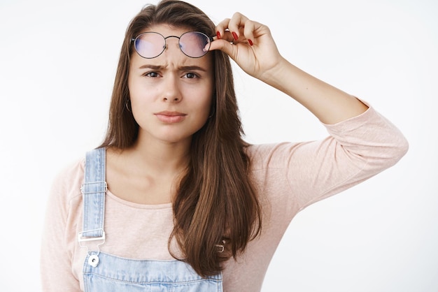 La ragazza che strizza gli occhi e accigliata perché non riesce a leggere il segno che ha una cattiva vista togliersi gli occhiali non può sembrare chiara mentre osserva l'immagine sfocata in piedi infastidita e interrogata su uno sfondo grigio