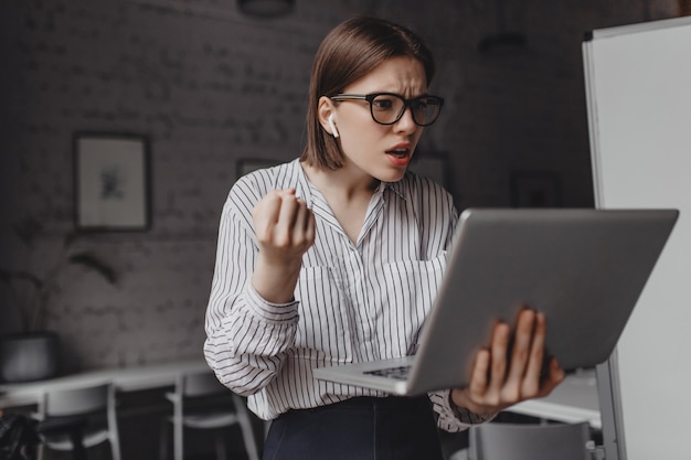 La ragazza comunica tramite video con indignazione. Donna in camicetta bianca e occhiali in posa con il computer portatile nel suo ufficio.