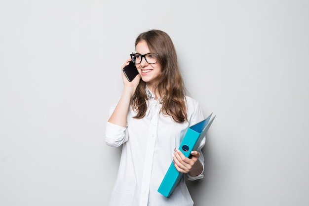 La ragazza con gli occhiali si è vestita in maglietta bianca dell'ufficio rigoroso sta davanti al muro bianco e tiene il suo telefono e la cartella nelle mani