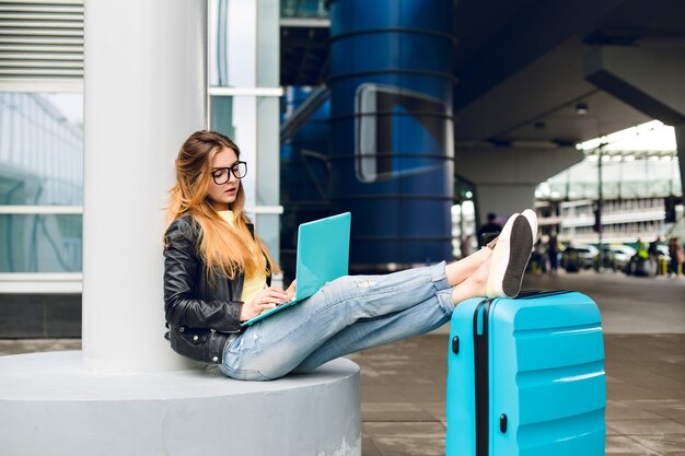 La ragazza con i capelli lunghi in occhiali neri è seduta fuori in aeroporto. Indossa jeans, giacca nera, scarpe gialle. Ha messo le gambe sulla valigia vicino. È annoiata a digitare sul laptop.