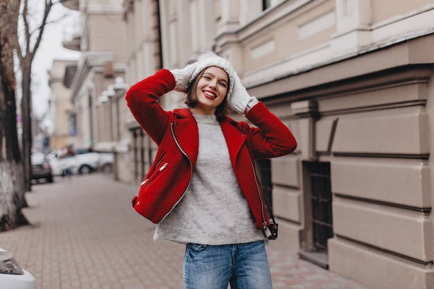 La ragazza con il rossetto rosso ride e mette il cappello lavorato a maglia. La donna in jeans e cappotto alla moda cammina nella città di autunno.