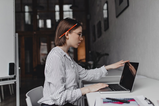 La ragazza con la matita rossa dietro l'orecchio lavora intensamente nel computer portatile mentre era seduto in un ampio ufficio bianco.