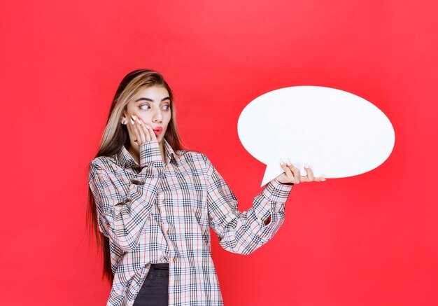 La ragazza con un maglione a quadri che tiene in mano un'ideaboard ovale sembra confusa ed eccitata