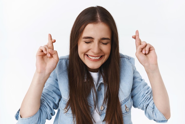 La ragazza crede di poter raggiungere l'obiettivo avendo fede sperando in attesa di risultati importanti incrociare le dita buona fortuna voler vincere sorridendo felicemente con gli occhi chiusi pregando in piedi sfondo bianco