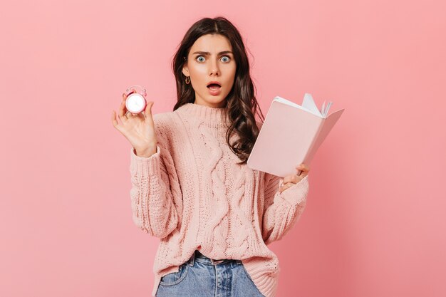 La ragazza dagli occhi azzurri in stato di shock esamina la fotocamera su sfondo rosa. Signora in maglione alla moda in posa con sveglia e diario.