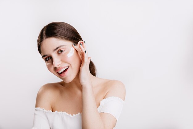 La ragazza dagli occhi verdi sorridente mette la crema sul viso pulito. Bruna in top bianco in posa sulla parete isolata.