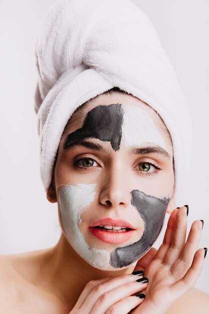 La ragazza dagli occhi verdi sta facendo la procedura facciale della stazione termale sulla parete bianca. Ritratto di donna in asciugamano con maschera all'argilla.