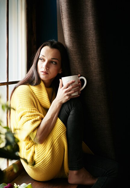 La ragazza dai capelli neri concepita sta guardando fuori dalla finestra e sta bevendo il tè