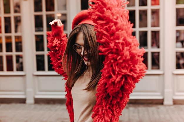 La ragazza dai capelli scuri attiva con gli occhiali si diverte per strada La donna con un elegante top beige e un cappotto rosso balla contro le finestre in cornici di legno bianco