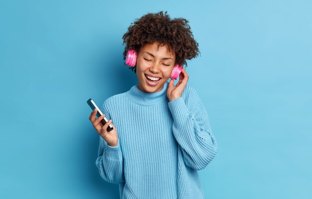 La ragazza dalla pelle scura indossa le danze delle cuffie durante la festa techno tiene lo smartphone chiude gli occhi con sorrisi di piacere indossa ampiamente un maglione casual