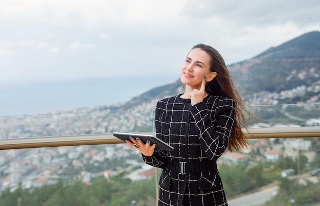 La ragazza del blogger del computer di Planshet sta pensando tenendo l'indice sulla guancia sullo sfondo della vista sulla città