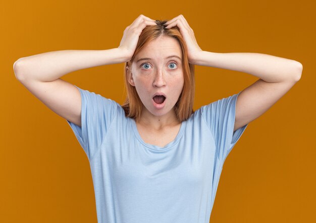 La ragazza dello zenzero giovane rossa scioccata con le lentiggini mette le mani sulla testa e guarda la telecamera sull'arancio