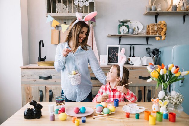 La ragazza felice che dipinge le uova per Pasqua vicino alla madre con il canestro