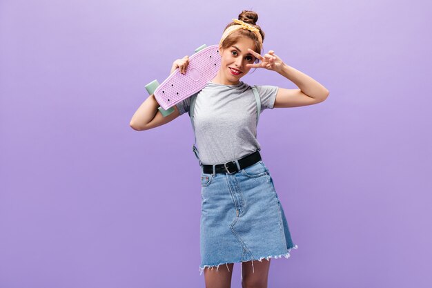 La ragazza felice in gonna di jeans mostra il segno di pace e tiene lo skateboard. Donna alla moda con bandana gialla in vestiti alla moda in posa.