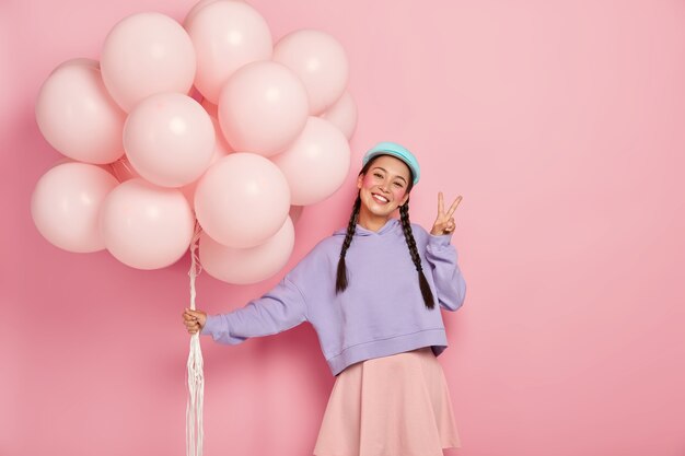 La ragazza felice saluta gli amici alla festa in mongolfiera, ha due trecce, indossa un maglione e una gonna viola, fa un gesto di pace, sta contro il muro rosa