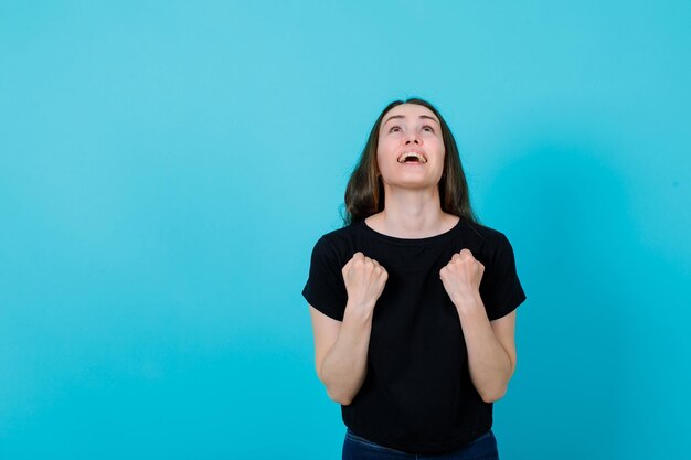 La ragazza felice sta cercando tenendo i pugni sul petto su sfondo blu