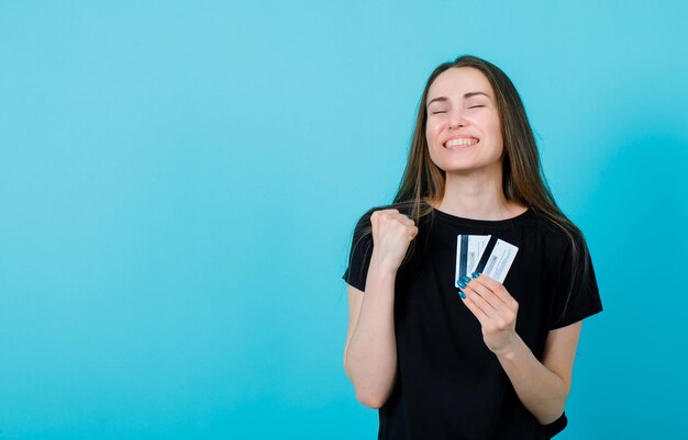 La ragazza felice sta desiderando alzando il pugno e tenendo le carte di credito su sfondo blu