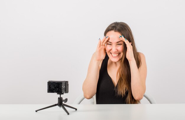 La ragazza felice sta esaminando la piccola macchina fotografica tenendo le mani sulla fronte su fondo bianco