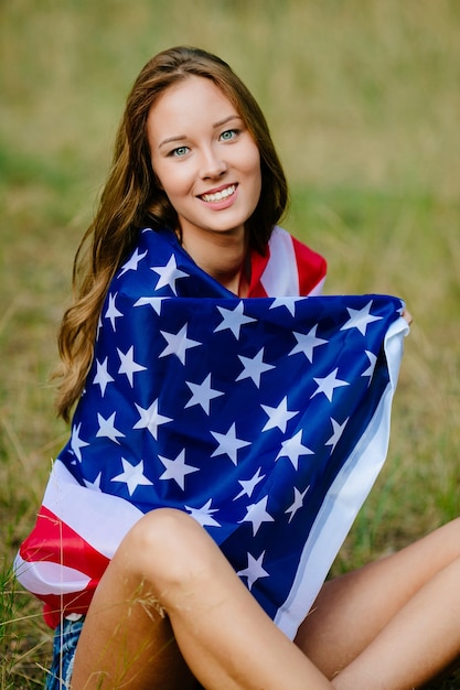 La ragazza felice sta sedendosi sull&#39;erba con la bandiera americana