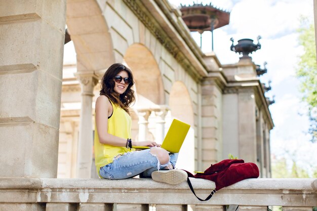 La ragazza graziosa del brunette sta sedendosi sul recinto sul vecchio fondo di architettura. Indossa una maglietta gialla, occhiali da sole, tiene un laptop giallo sulle ginocchia, sorride alla telecamera.