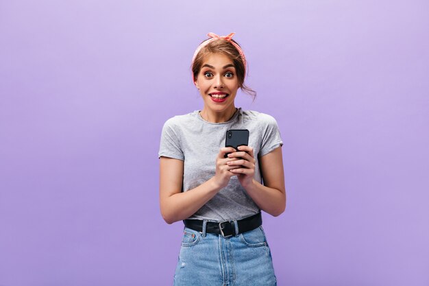 La ragazza guarda felicemente e tiene lo smartphone. Giovane donna sorpresa in maglietta grigia e gonna moderna che esamina la macchina fotografica su sfondo viola.