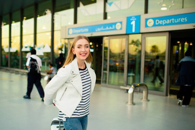 La ragazza in giacca bianca e camicia striata cammina con valigia dall&#39;aeroporto