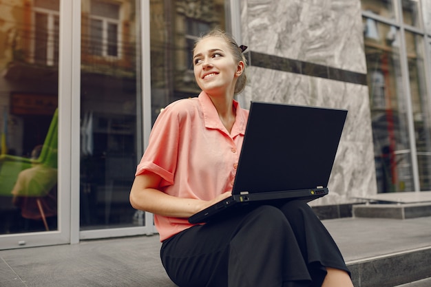 La ragazza in una camicia rosa che si siede vicino alla casa e usa il computer portatile