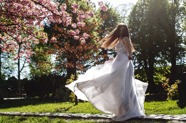 La ragazza in vestito bianco sta sotto l&#39;albero di sakura di fioritura rosa nel parco