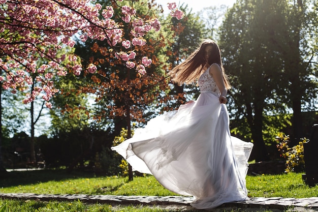 La ragazza in vestito bianco sta sotto l&#39;albero di sakura di fioritura rosa nel parco