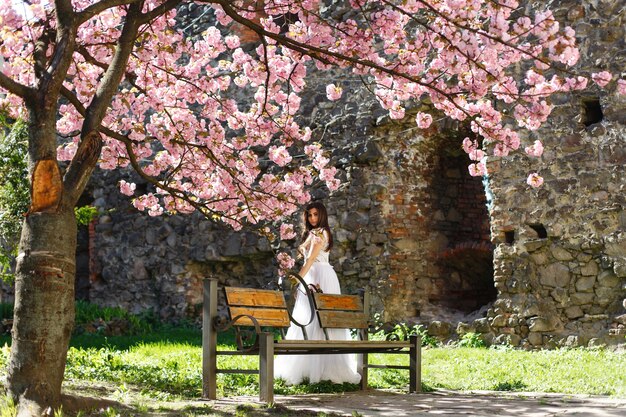 La ragazza in vestito bianco sta sotto l&#39;albero di sakura di fioritura rosa nel parco