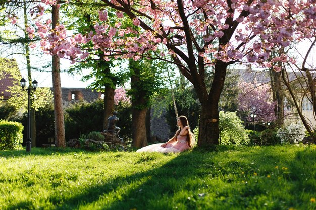 La ragazza in vestito rosa si siede sotto l&#39;albero di sakura di fioritura nel parco