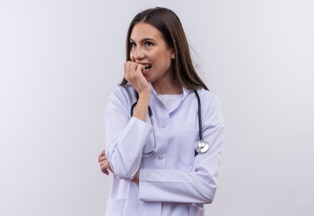 La ragazza interessata che indossa l'abito medico dello stetoscopio mise la mano sul mento sulla parete bianca isolata