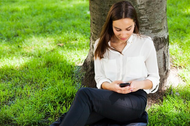 La ragazza lavora con il suo telefono seduto sotto l&#39;albero