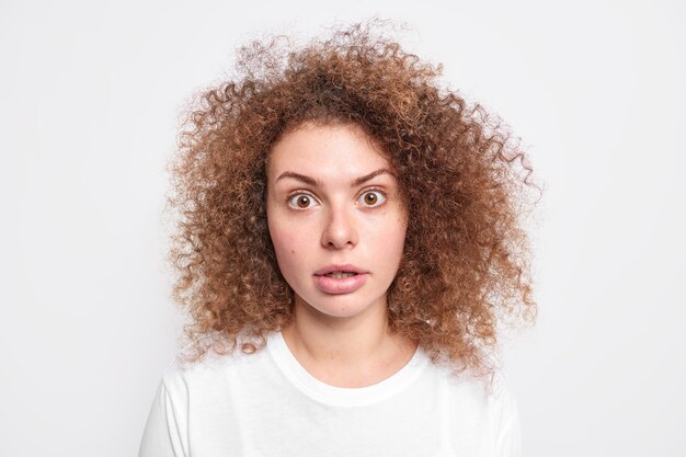 La ragazza millenaria scioccata con i capelli ricci e folti ha dimenticato qualcosa colpito da una rivelazione sorprendente, gli occhi sono spuntati fuori vestita con una maglietta casual isolata sul muro bianco. Concetto di reazione