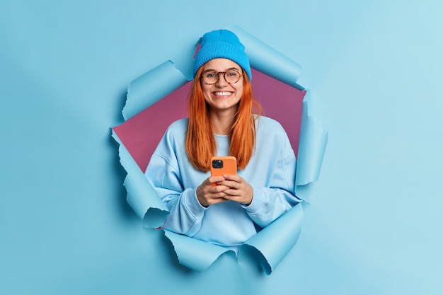 La ragazza millenaria sorridente felice con i capelli rossi tiene il cellulare moderno gode di mandare SMS nei social media utilizza i servizi di rete mobile indossa un maglione e un cappello blu.