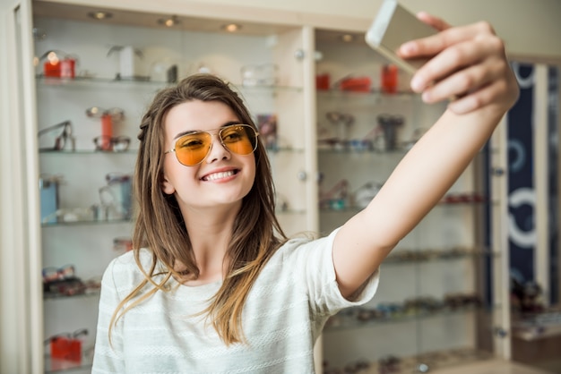 La ragazza non vive mai a casa senza smartphone, elegante bruna europea che prova gli occhiali da sole mentre fa selfie sul telefono, sorridendo ampiamente