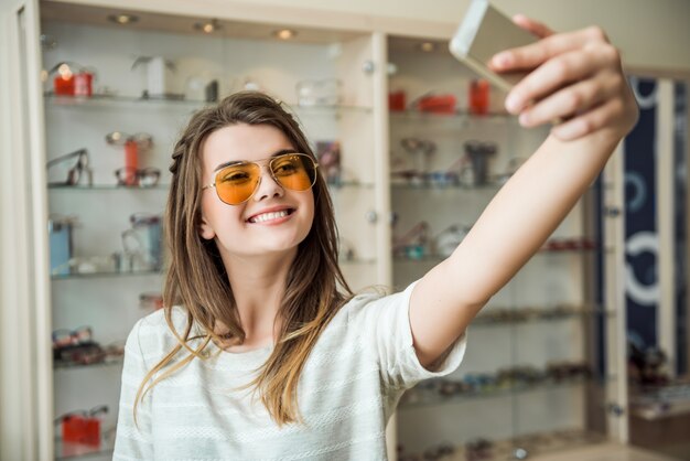 La ragazza non vive mai a casa senza smartphone, elegante bruna europea che prova gli occhiali da sole mentre fa selfie sul telefono, sorridendo ampiamente