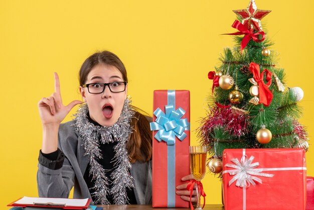 La ragazza perplessa di vista frontale con gli occhiali che si siedono al tavolo ha indicato con il dito sull'albero di Natale e sul cocktail dei regali