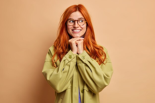 La ragazza positiva allo zenzero di bell'aspetto sorride piacevolmente tiene le mani sotto il mento e distoglie lo sguardo vestito con una giacca verde esprime emozioni positive.