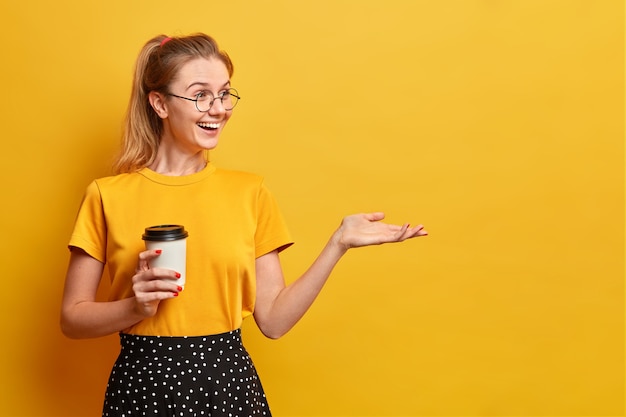 La ragazza positiva sincera beve caffè aromatico dalla tazza usa e getta tiene la mano alzata