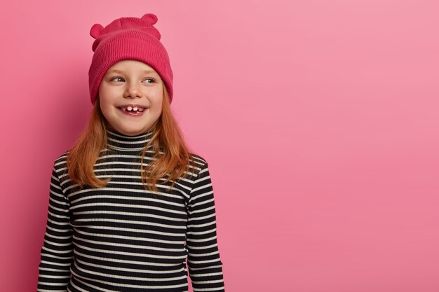 La ragazza rossa positiva indossa cappello e poloneck a strisce, guarda da parte, nota qualcosa di divertito, ricorda di aver passato del tempo piacevole con i genitori, posa su un muro rosa pastello, ha un'espressione adorabile