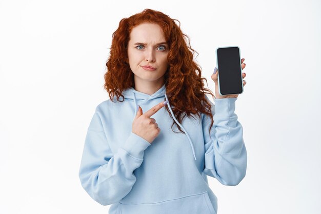 La ragazza rossa scettica sembra confusa, indicando lo schermo vuoto del telefono cellulare con la faccia dubbiosa, in piedi sconvolta con l'app su bianco