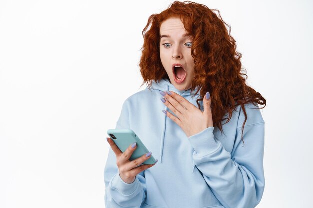 La ragazza rossa scioccata e spaventata urla mentre legge il messaggio sullo smartphone, fissando lo schermo del cellulare con la faccia sorpresa, in piedi sul bianco